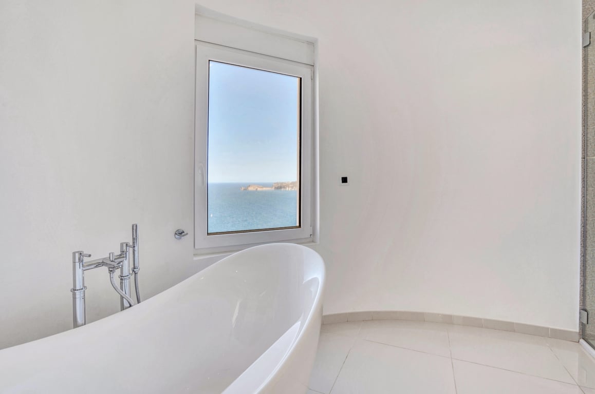 Minimalistisches Badezimmer mit freistehender weißer Badewanne und Chromarmaturen, mit direktem Blick auf das Mittelmeer durch das Fenster.
