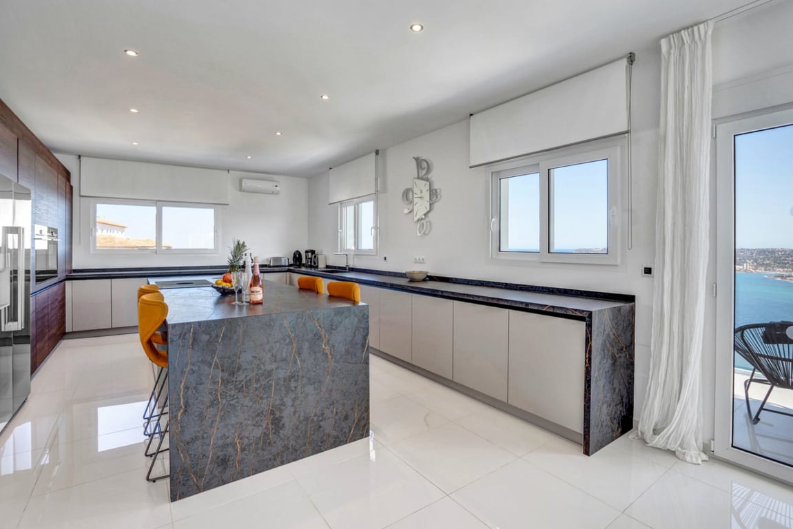 Moderne Designerküche mit dunkler Steininsel, senfgelben Hockern und integrierten Geräten. Große Glasfronten ermöglichen einen weiten Blick über die Bucht von Jávea.