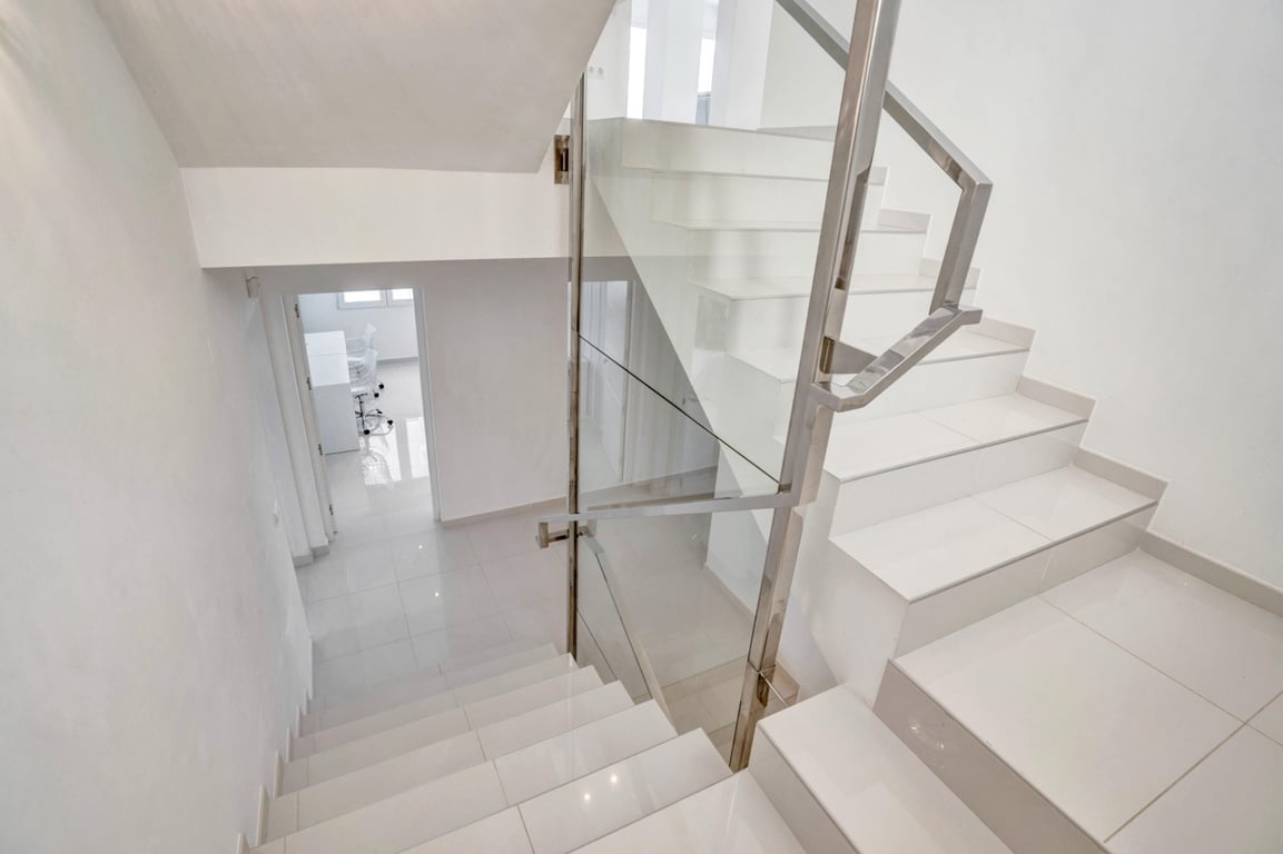 Moderne Treppe mit weißen Fliesenstufen, Glaspaneelen und Edelstahlgeländern, die ein helles und minimalistisches Innendesign unterstreicht.
