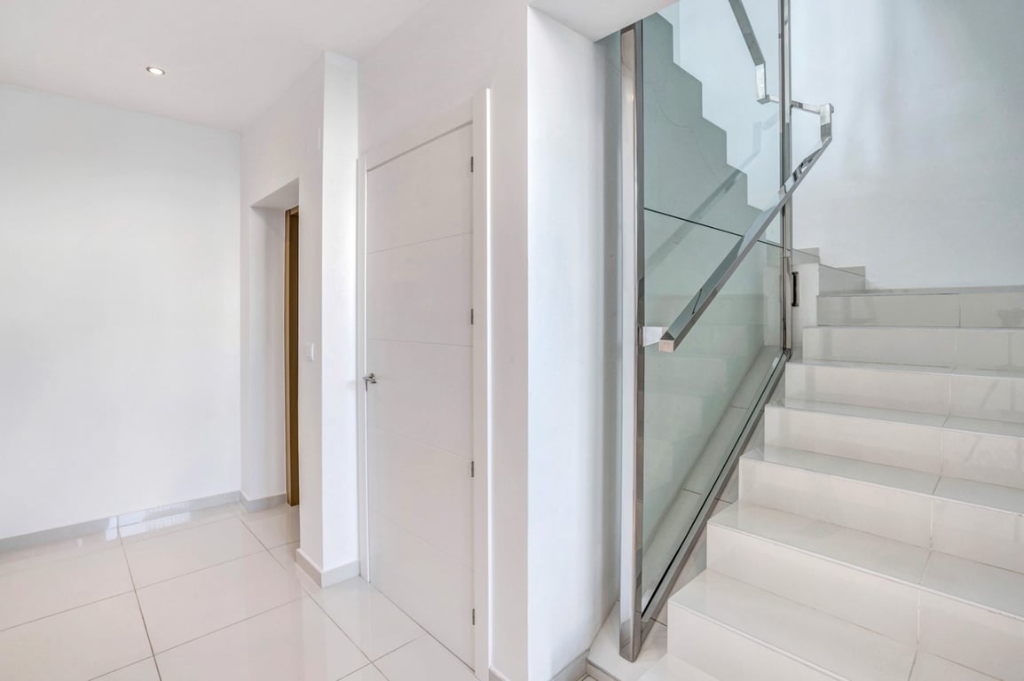 Moderner Flur mit weißen Bodenfliesen und einer Treppe mit Glasgeländer und Handläufen aus Edelstahl.