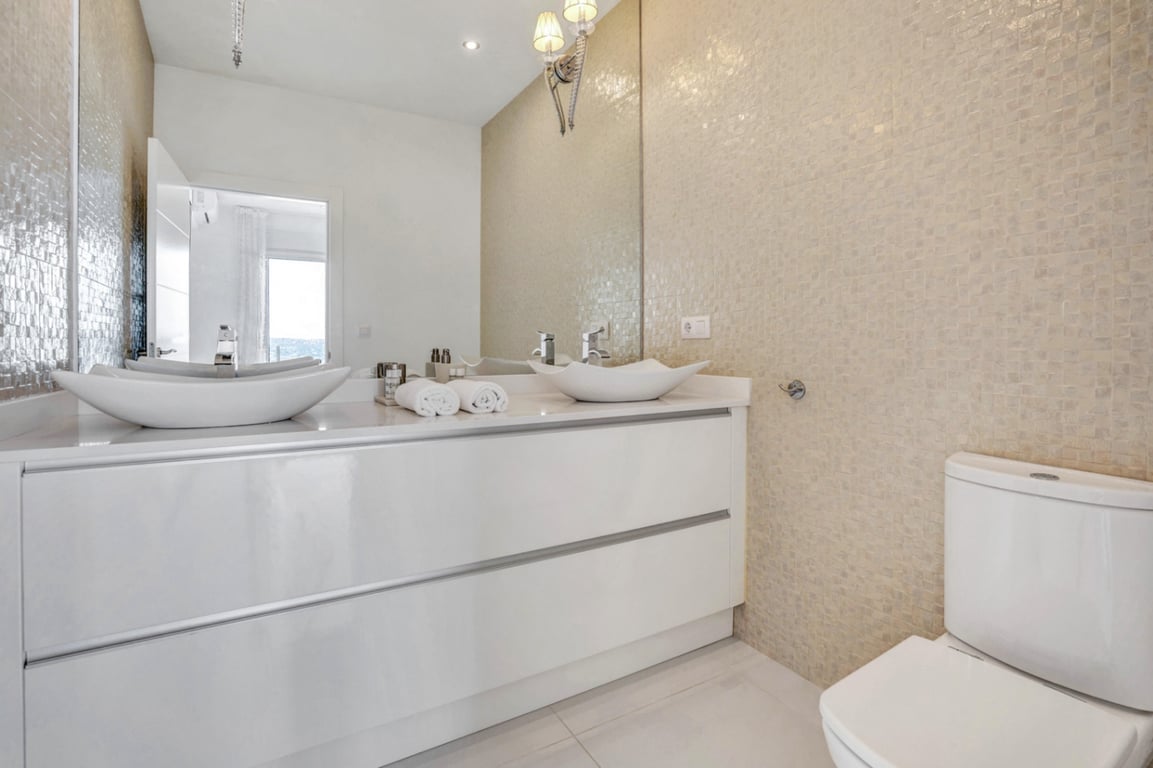 Modernes Badezimmer mit Doppelwaschtisch, glänzend weißen Unterschränken, Aufsatzbecken und einer Wand aus beigem Mosaik.