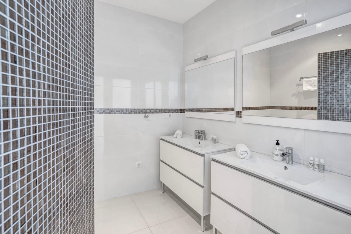 Modernes Badezimmer mit Doppelwaschtisch, minimalistischen weißen Unterschränken und dekorativen dunklen Mosaikfliesen an den Wänden.