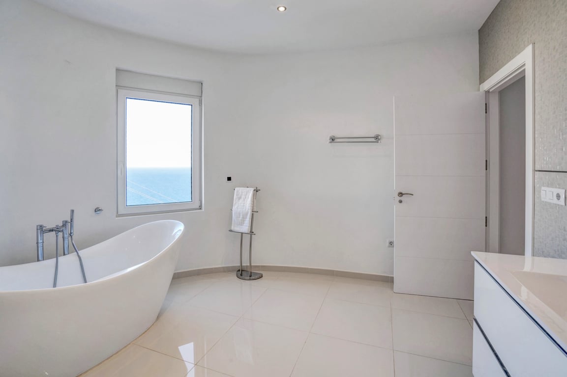 Modernes Badezimmer mit freistehender Badewanne, hellen Fliesenböden und Fenster mit direktem Blick auf das Mittelmeer.