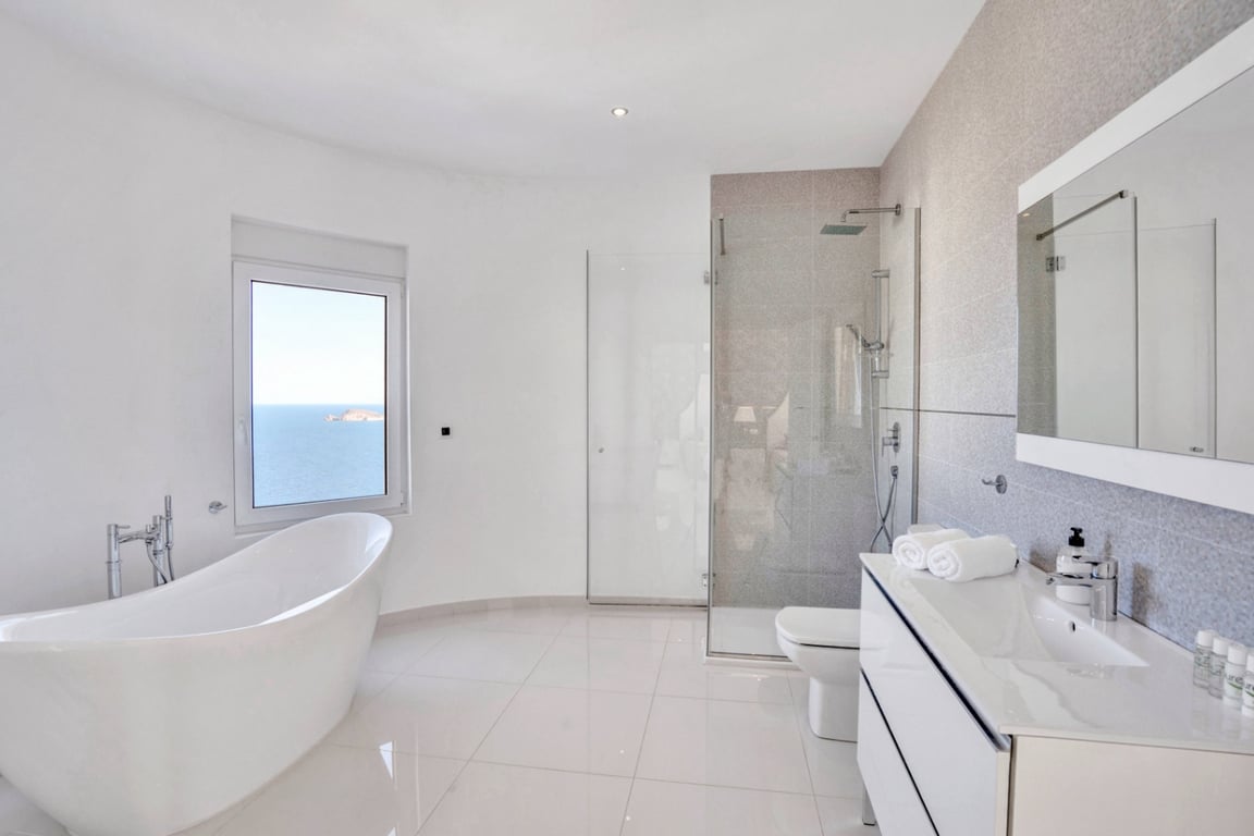 Modernes Badezimmer mit freistehender Badewanne und Meerblick, einer begehbaren Glasdusche sowie einem weißen Waschtisch mit großem Spiegel.