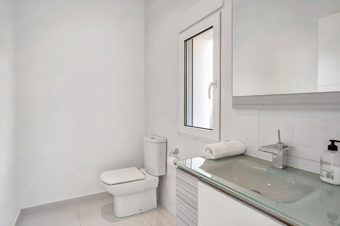 Modernes Badezimmer mit lichtdurchlässigem Glaswaschtisch, verchromten Armaturen und weiß gefliesten Wänden mit kleinem Fenster.