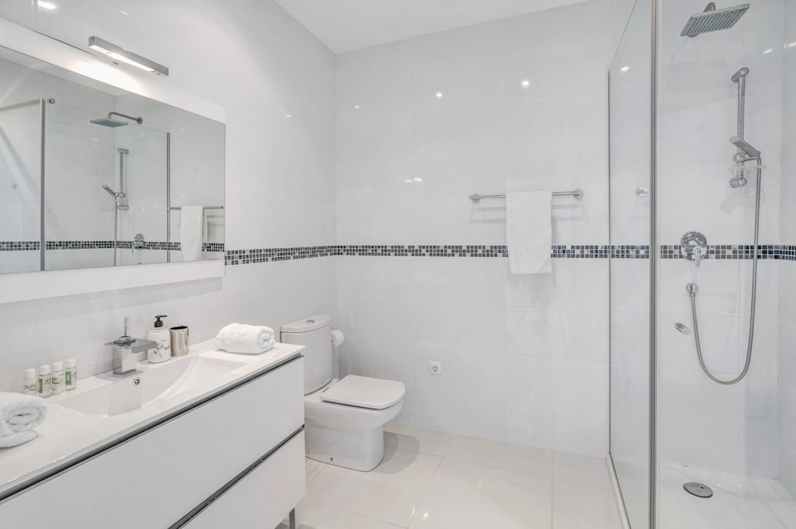 Modernes Badezimmer mit weißen Fliesen und Mosaikbordüre, einem weißen Waschtisch, WC und einer begehbaren Glasdusche mit verchromten Armaturen.