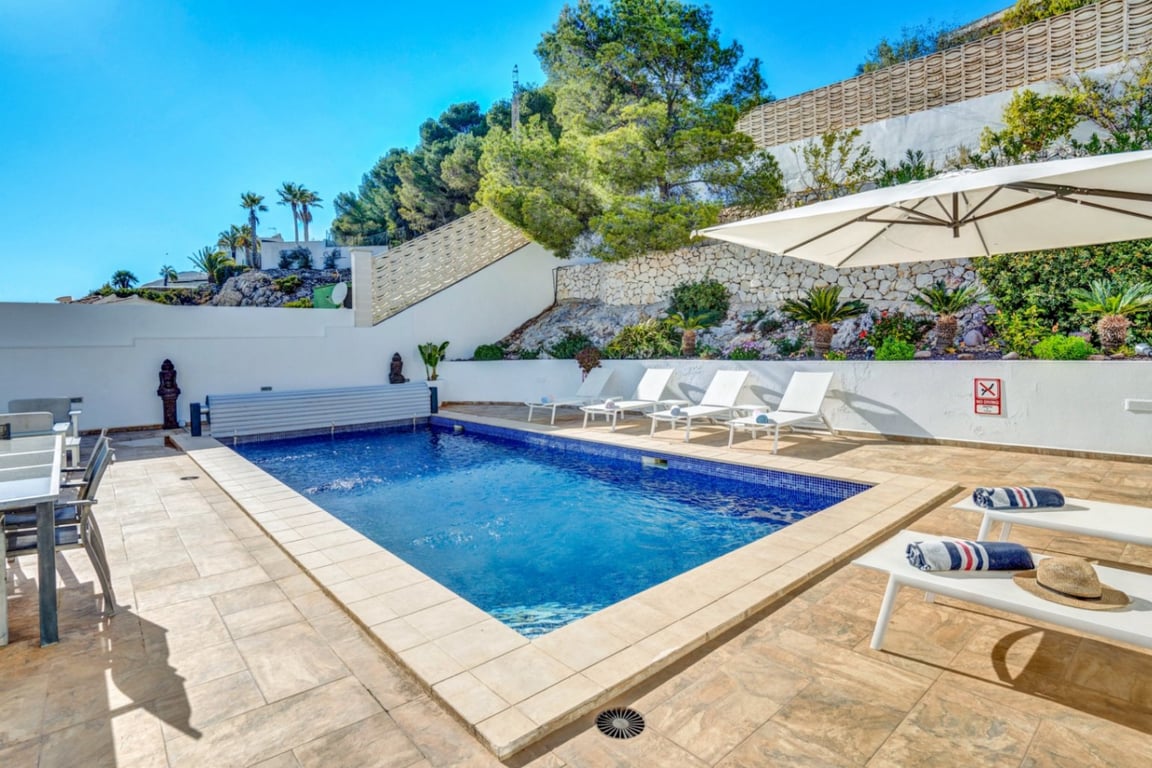 Privater Außenpool mit blauem Mosaik, großzügiger Sonnenterrasse, weißen Liegen und einer Natursteinmauer inmitten mediterraner Bepflanzung.