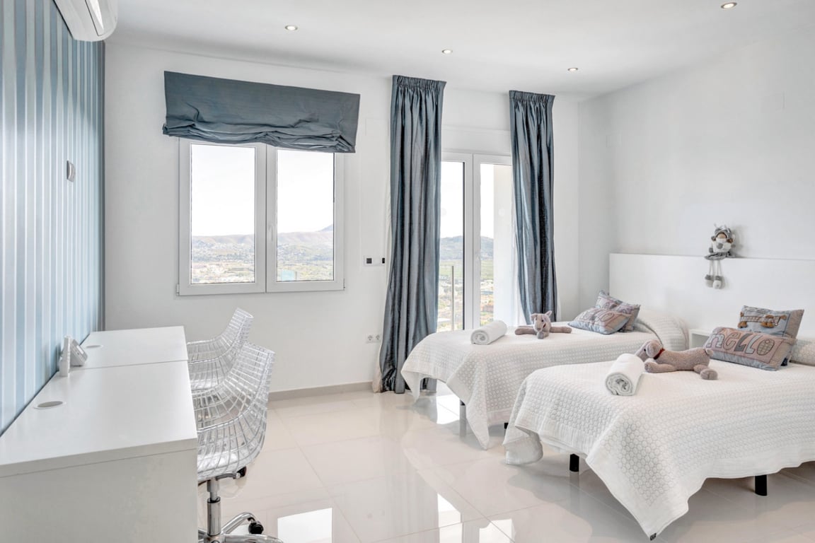 Schlafzimmer mit zwei Einzelbetten, weißem Schreibtisch und Designstühlen sowie großen Fenstern mit Panoramablick auf Berge und Meer.