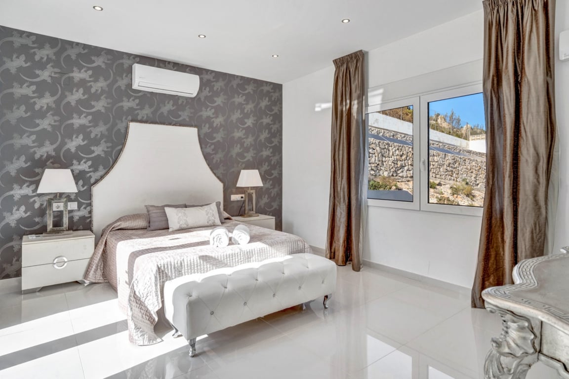 Schlafzimmer-Suite mit grauer Mustertapete, weißem Fliesenboden, Klimaanlage und Fenster mit Blick auf die charakteristische Felswand des Grundstücks.
