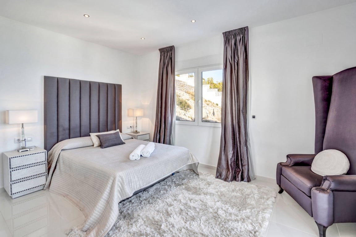 Schlafzimmer-Suite mit hohem grauem Kopfteil, flauschigem weißem Teppich und markantem Ohrensessel. Einbaustrahler und Fenster mit Blick auf die Felsformationen.