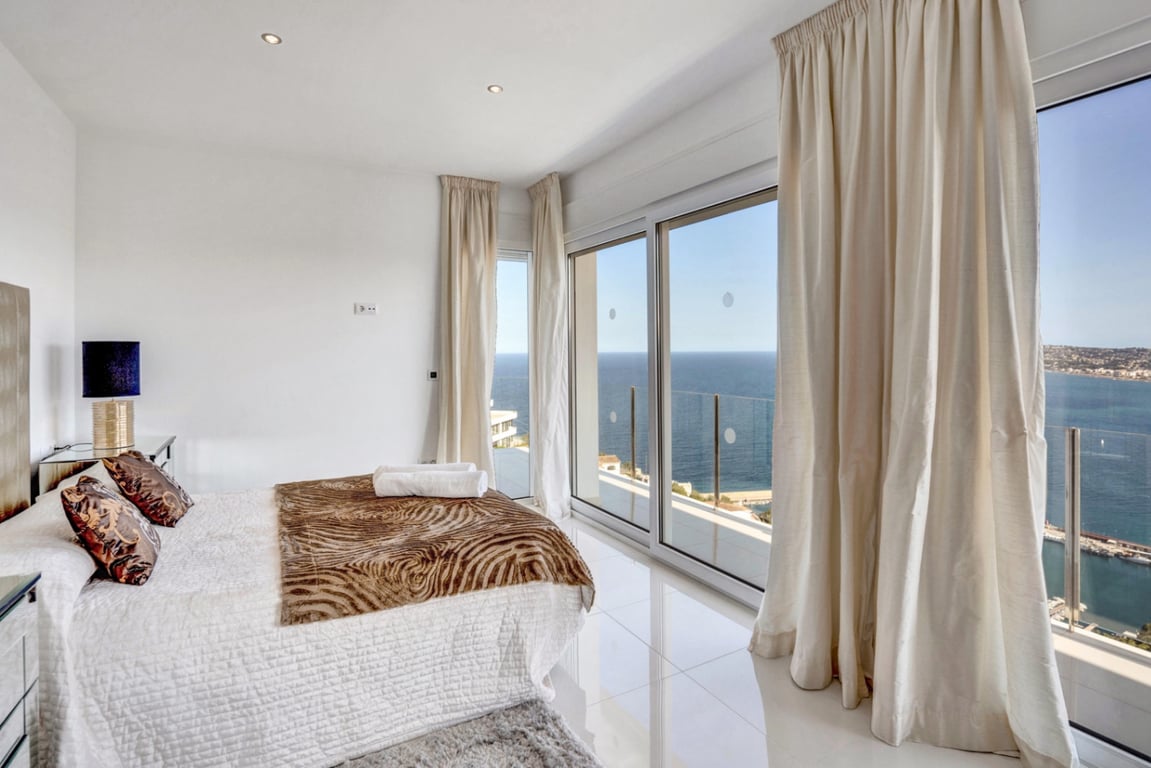 Schlafzimmer-Suite mit weißem Hochglanzboden, bodentiefen Glasschiebetüren und direktem Zugang zum Balkon mit Panoramablick auf das Mittelmeer und den Hafen von Jávea.