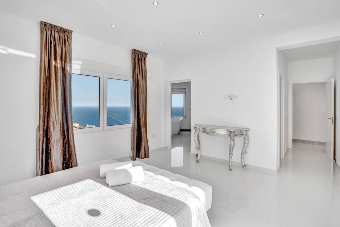 Schlafzimmer-Suite mit weißen Hochglanzfliesen, Meerblick und angrenzendem Badezimmer mit freistehender Badewanne und Panoramafenster.