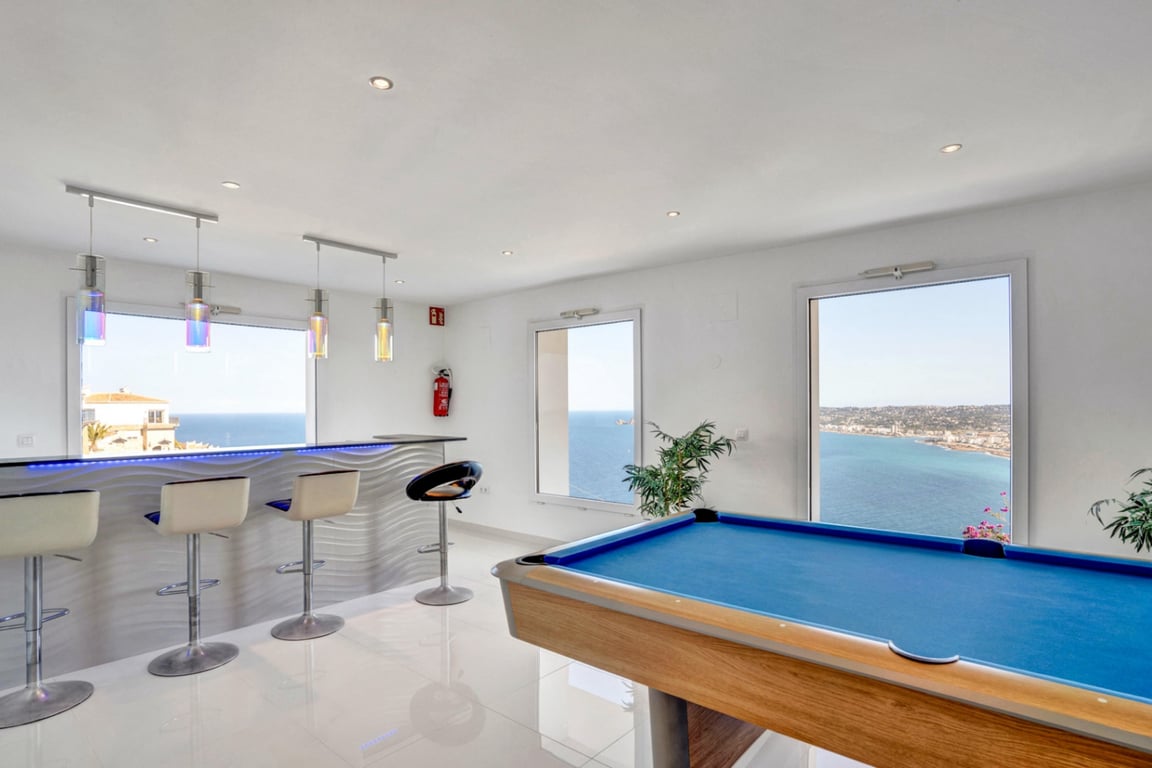 Spielzimmer mit blauem Billardtisch und moderner Bar mit LED-Beleuchtung, die durch große Fenster einen Panoramablick auf das Meer bietet.