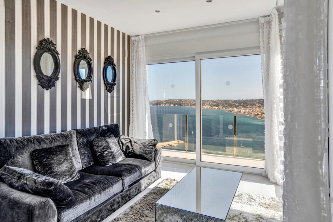 Wohnbereich mit grauem Samtsofa, gestreifter Tapete und verzierten Spiegeln, ergänzt durch bodentiefe Fenster mit weitem Blick auf das Meer.