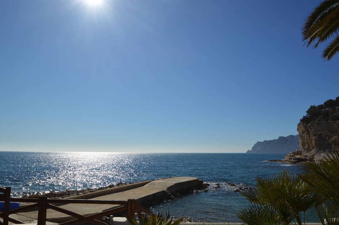 Blick auf die Cala Advocat mit einem Betonsteg und glitzerndem Mittelmeerwasser. Die Szene zeigt felsige Klippen und mediterrane Palmen bei Sonnenschein.