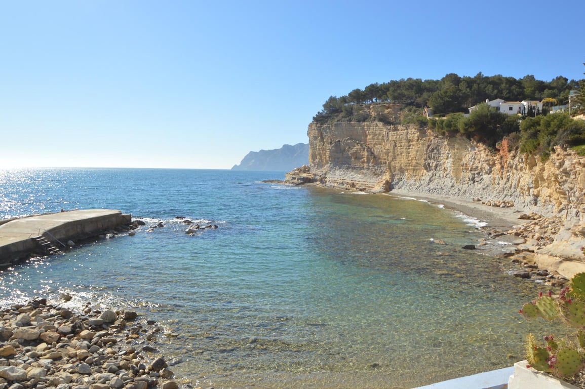 Blick auf die Cala Advocat mit klarem, türkisblauem Wasser, einer kleinen Betonmole und markanten Kalksteinfelsen unter strahlend blauem Himmel.