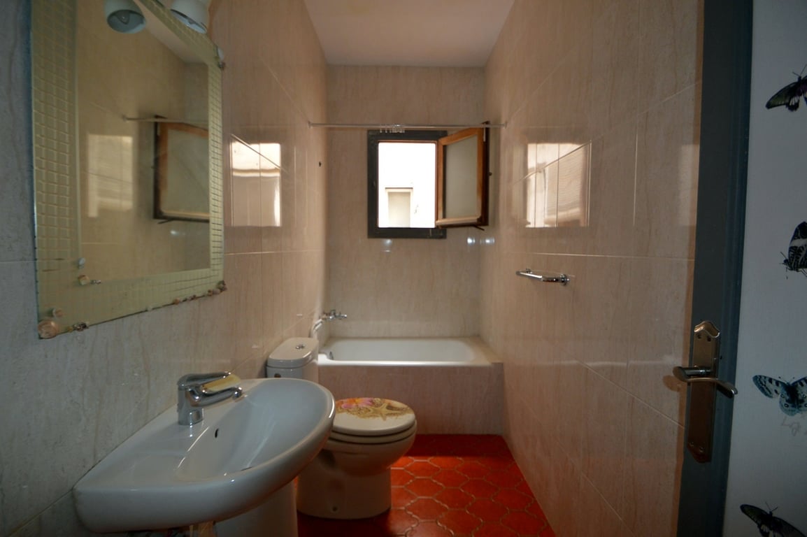 Badezimmer mit roten sechseckigen Bodenfliesen, cremefarbenen Wandfliesen, einem Standwaschbecken und einer Einbaubadewanne unter einem kleinen Fenster zur Belüftung.