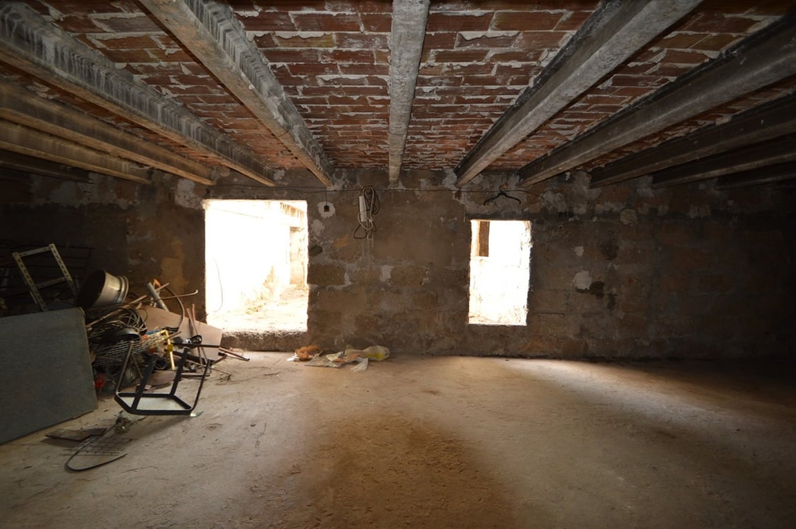 Großzügiger, renovierungsbedürftiger Innenraum mit Betonträgern und Sichtziegeldecke. Der Raum bietet Natursteinwände und zwei Lichtöffnungen nach draußen.