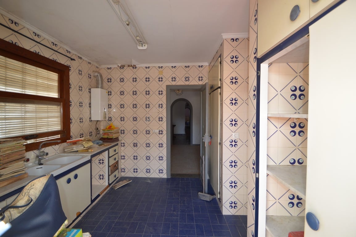 Küche mit gemusterten Wandfliesen, blauem Fliesenboden und weißen Schränken mit blauen Griffen. Sie verfügt über ein Doppelspülbecken am Holzfenster und Zugang zum Innenflur.