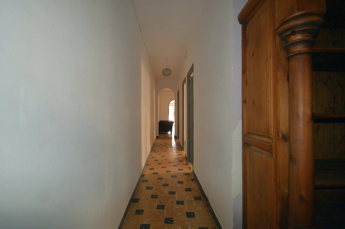 Langer Flur mit traditionell gemusterten Fliesen und weißen Wänden, der zu einem Rundbogen und einem massiven Holzschrank mit Schnitzereien führt.
