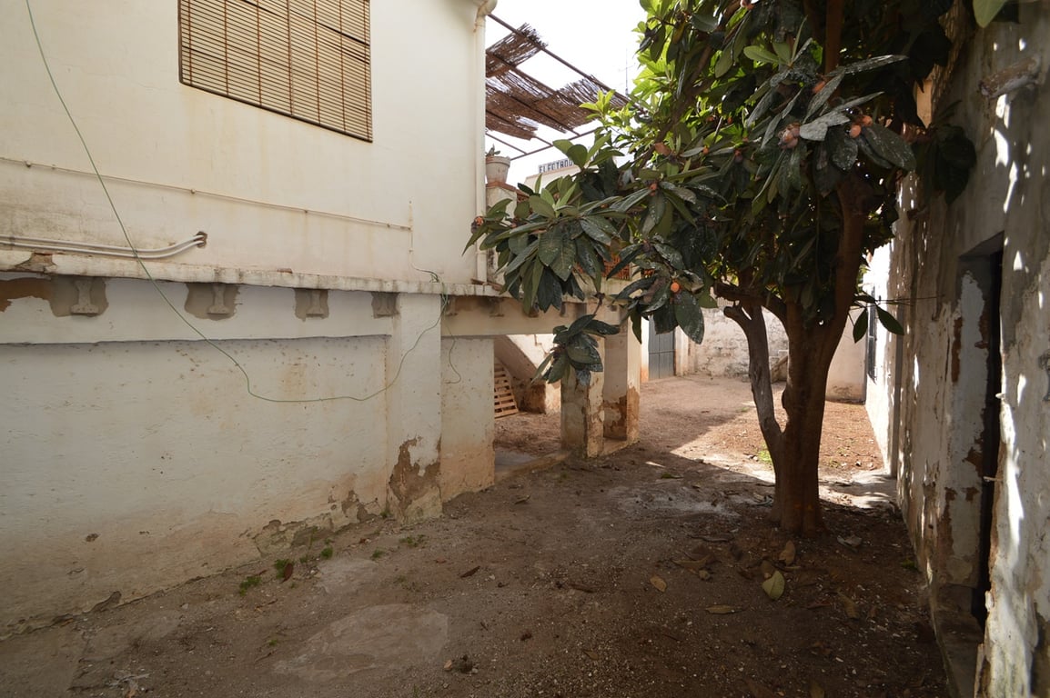 Privater Innenhof mit einem Mispelbaum, weiß getünchten Wänden und einem überdachten Bereich unter der Terrasse, ideal für ein Renovierungsprojekt.