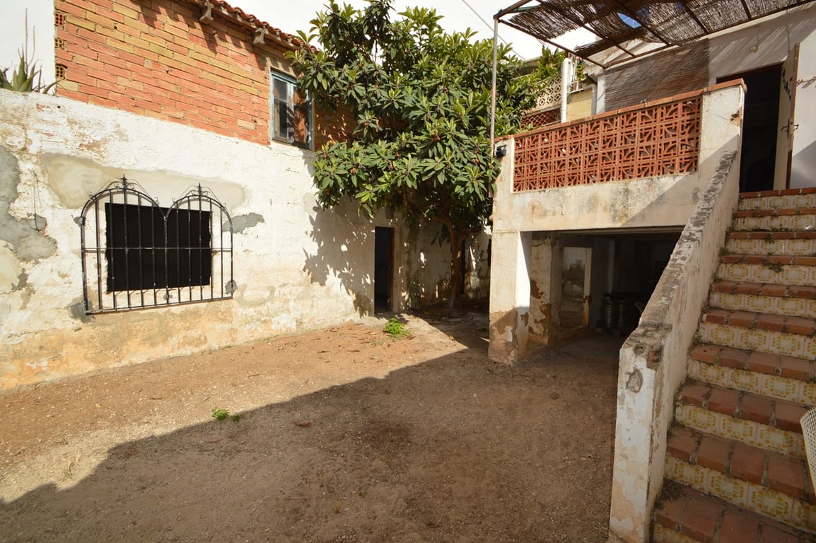 Rustikaler Innenhof mit Erdboden, großem Baum und Außentreppe aus Fliesen, die zu einer Terrasse mit Schilfrohr-Pergola und Keramikgeländer führt.