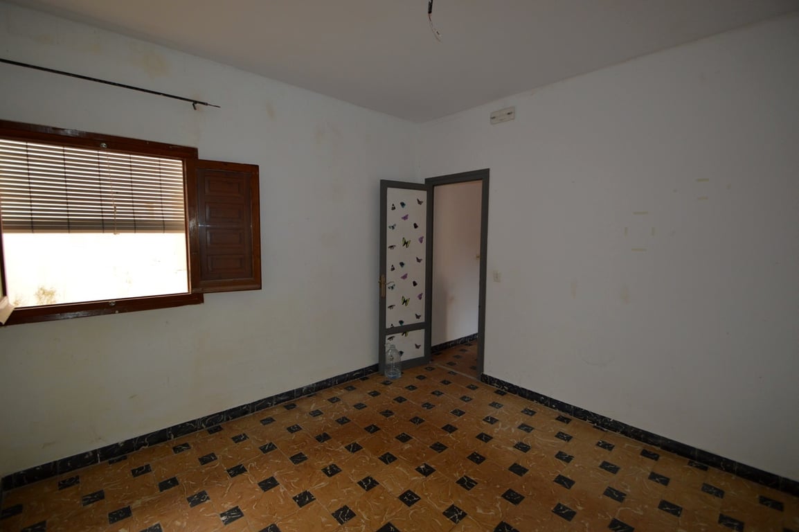 Schlafzimmer mit traditionell gemusterten Bodenfliesen, weißen Wänden und Holzfenster mit Läden. Ein renovierungsbedürftiger Raum mit Potenzial.