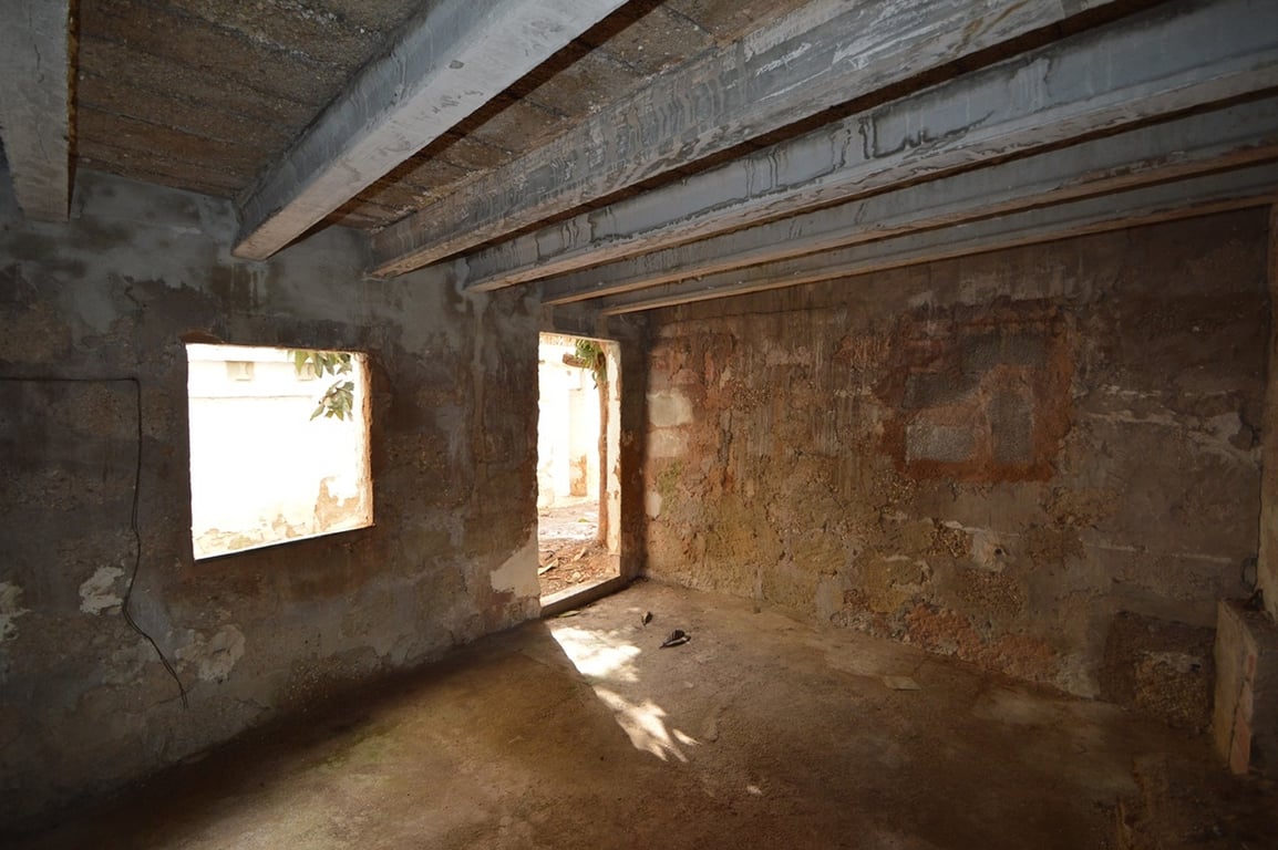 Unbehandelter Innenraum mit originalen Natursteinwänden und sichtbaren Deckenbalken. Natürliches Licht fällt durch eine Fensteröffnung und den Zugang zum Innenhof ein.