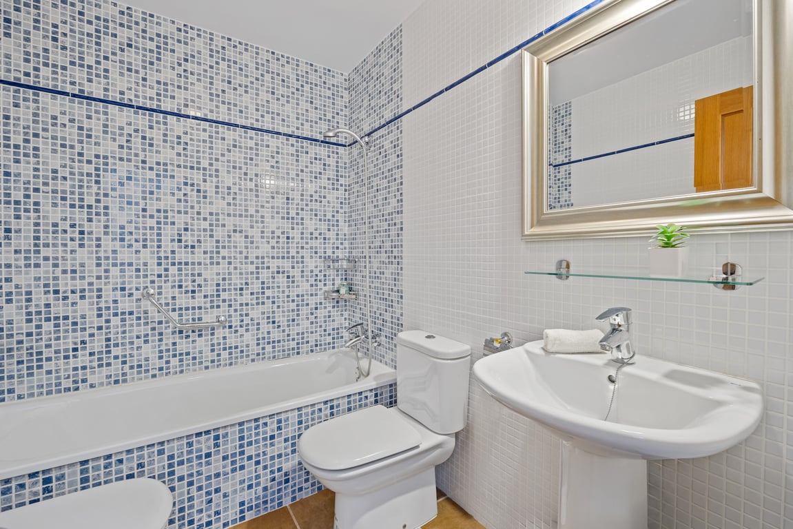 Funktionales Badezimmer mit Badewanne, blau-weißen Mosaikfliesen, Säulenwaschbecken und einem großen Spiegel mit silbernem Rahmen.