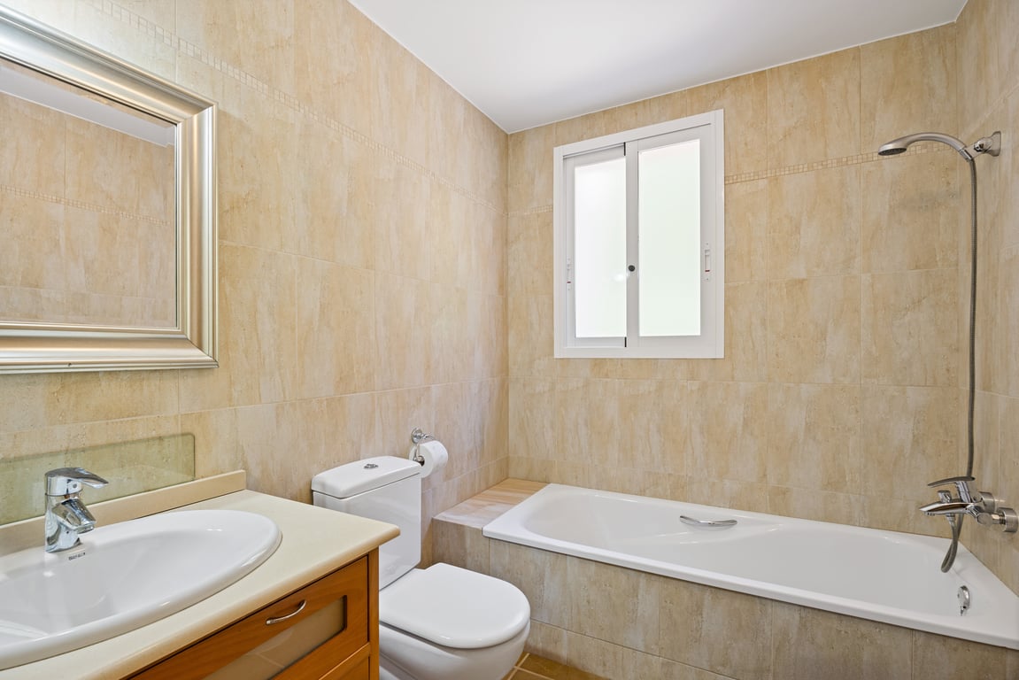 Funktionales Badezimmer mit beigen Wandfliesen, Einbaubadewanne mit Dusche, Holzwaschtisch und einem Milchglasfenster für natürliches Licht und Privatsphäre.