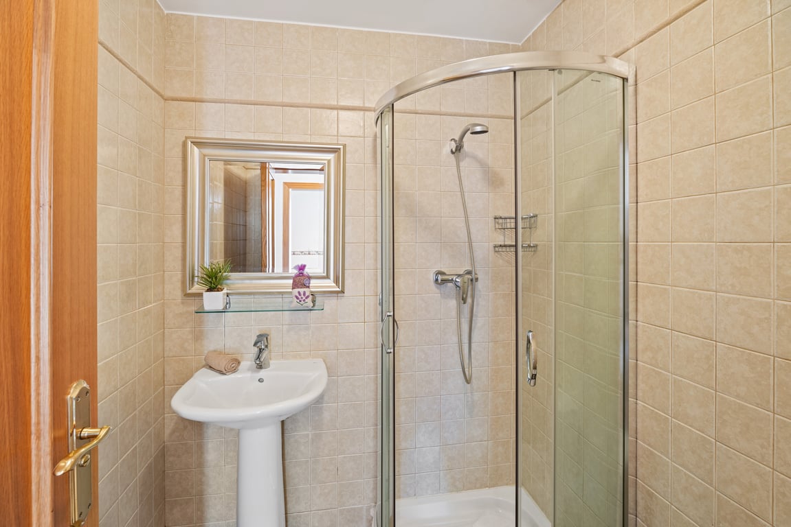 Funktionales Badezimmer mit einer abgerundeten Eckdusche aus Glas, einem weißen Säulenwaschbecken und beigefarbenen Keramikfliesen.
