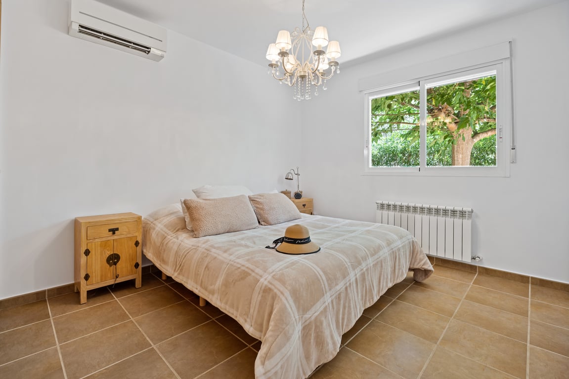 Helles Schlafzimmer mit Fliesenboden, Split-Klimaanlage und Zentralheizung. Ein Fenster bietet Ausblick ins Grüne, ergänzt durch einen dekorativen Kronleuchter.