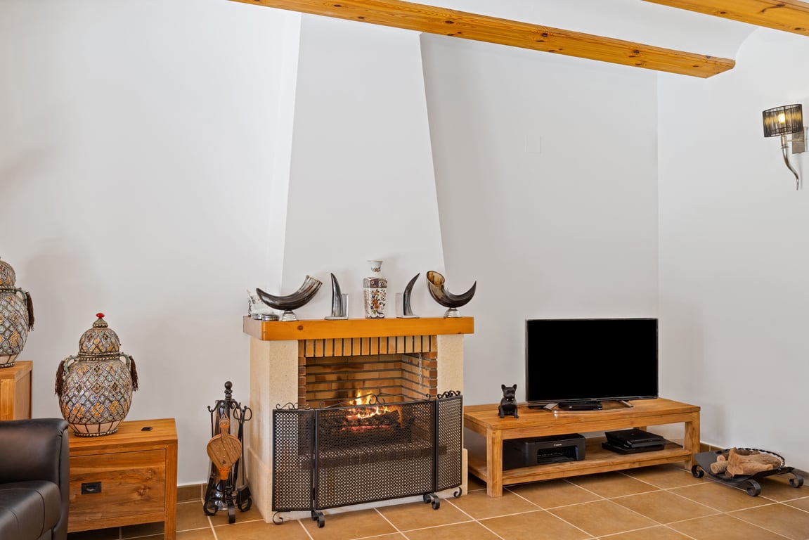 Wohnbereich mit traditionellem Kamin, sichtbaren Holzbalken an der Decke und rustikalen Bodenfliesen. Holzmöbel und Dekoration sorgen für ein gemütliches Ambiente.