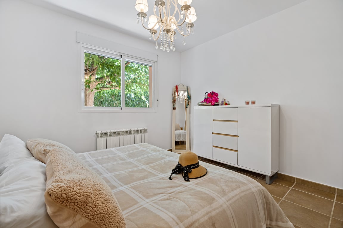 Helles Schlafzimmer mit weißen Wänden, Fliesenboden, moderner Kommode, klassischem Kristallleuchter und Fenster mit Blick ins Grüne.
