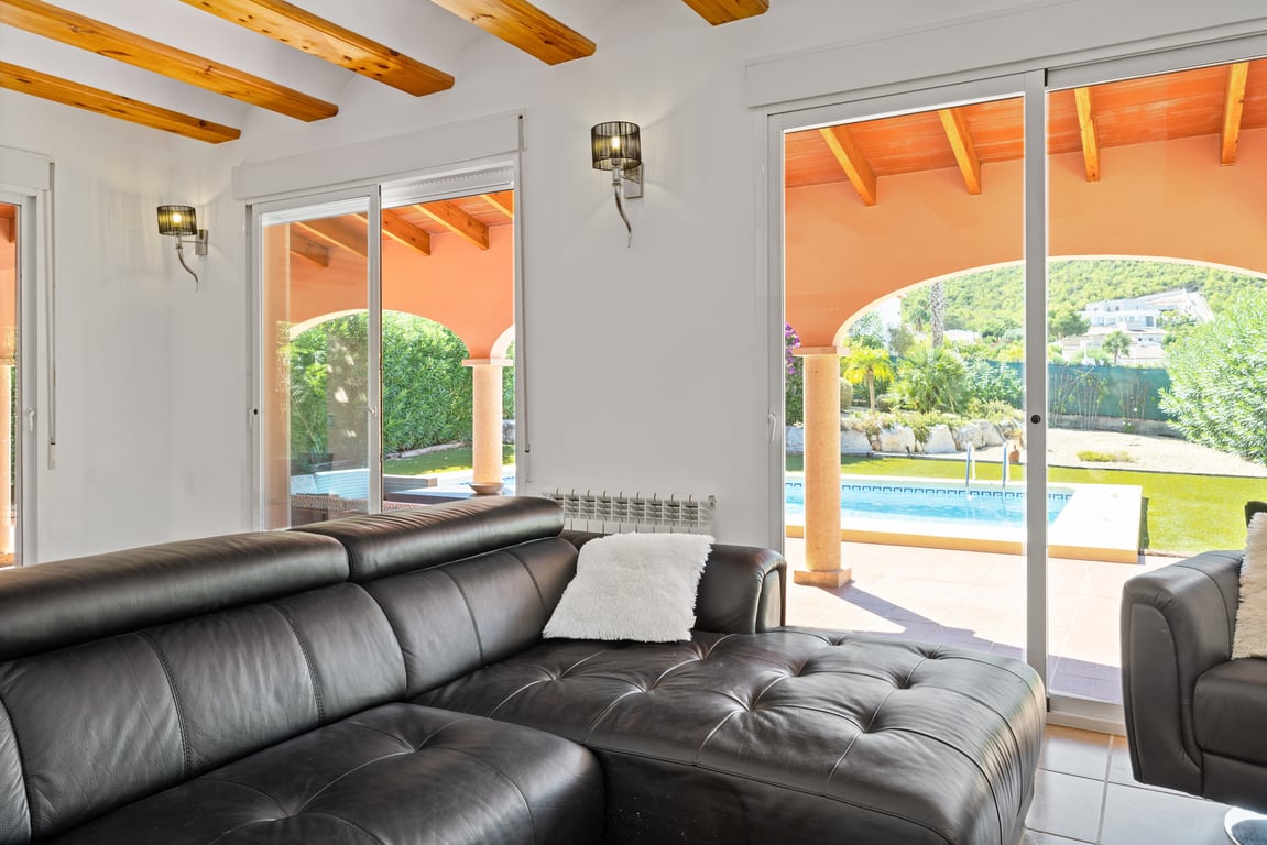 Wohnzimmer mit sichtbaren Holzbalken und schwarzem Ledersofa, das durch große Glasschiebetüren direkten Zugang zur Naya und zum Poolbereich bietet.