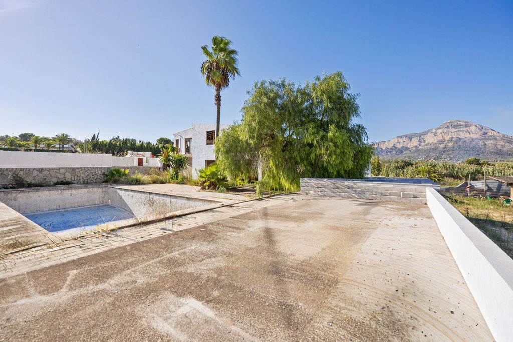 Außen-Betonterrasse mit Panoramablick auf den Montgó, renovierungsbedürftigem Pool und einem Gebäude im mediterranen Stil mit Palmen.