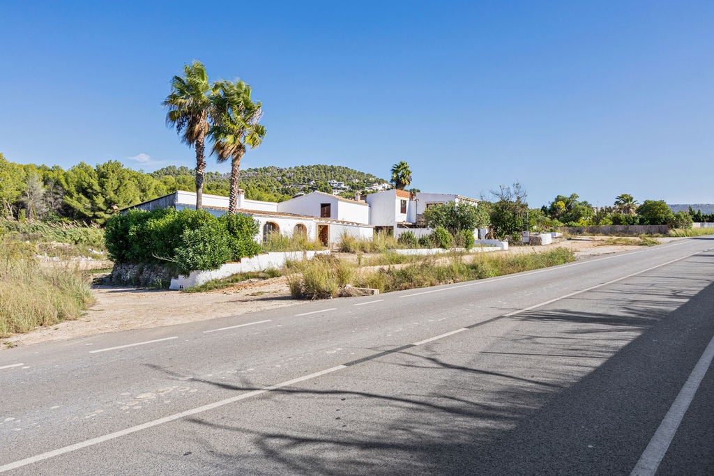 Außenansicht des mediterranen Anwesens von der Straße aus, mit weißen Fassaden, Palmen und bewaldeten Hügeln unter klarem Himmel.