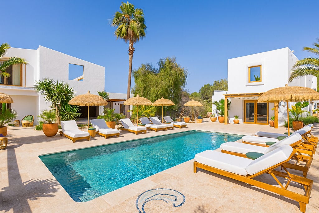 Außenpoolbereich mit Natursteinpflaster, Sonnenliegen aus Holz mit weißen Auflagen und Reet-Sonnenschirmen vor weißer mediterraner Architektur.