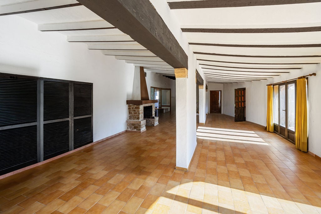 Großzügiger, offener Raum mit dunklen Holzbalkendecken, Terrakottaböden und einem rustikalen Steinkamin. Große Fensterfronten sorgen für viel Tageslicht.