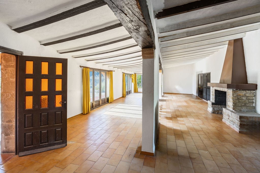 Großzügiger rustikaler Saal mit sichtbaren dunklen Holzbalken, traditionellem Terrakottaboden und einem Natursteinkamin mit Metallhaube.