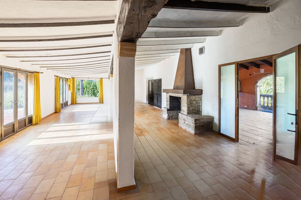 Großzügiger rustikaler Wohnbereich mit Terrakottaboden, sichtbaren Holzbalken und einem markanten Steinkamin. Große Fensterfronten sorgen für helles, natürliches Licht.