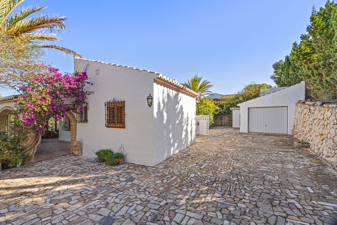 Außenbereich mit unregelmäßigem Steinpflaster, freistehender Garage und weißer Fassade, geschmückt mit rosa Bougainvillea und traditionellen Tosca-Steinbögen.