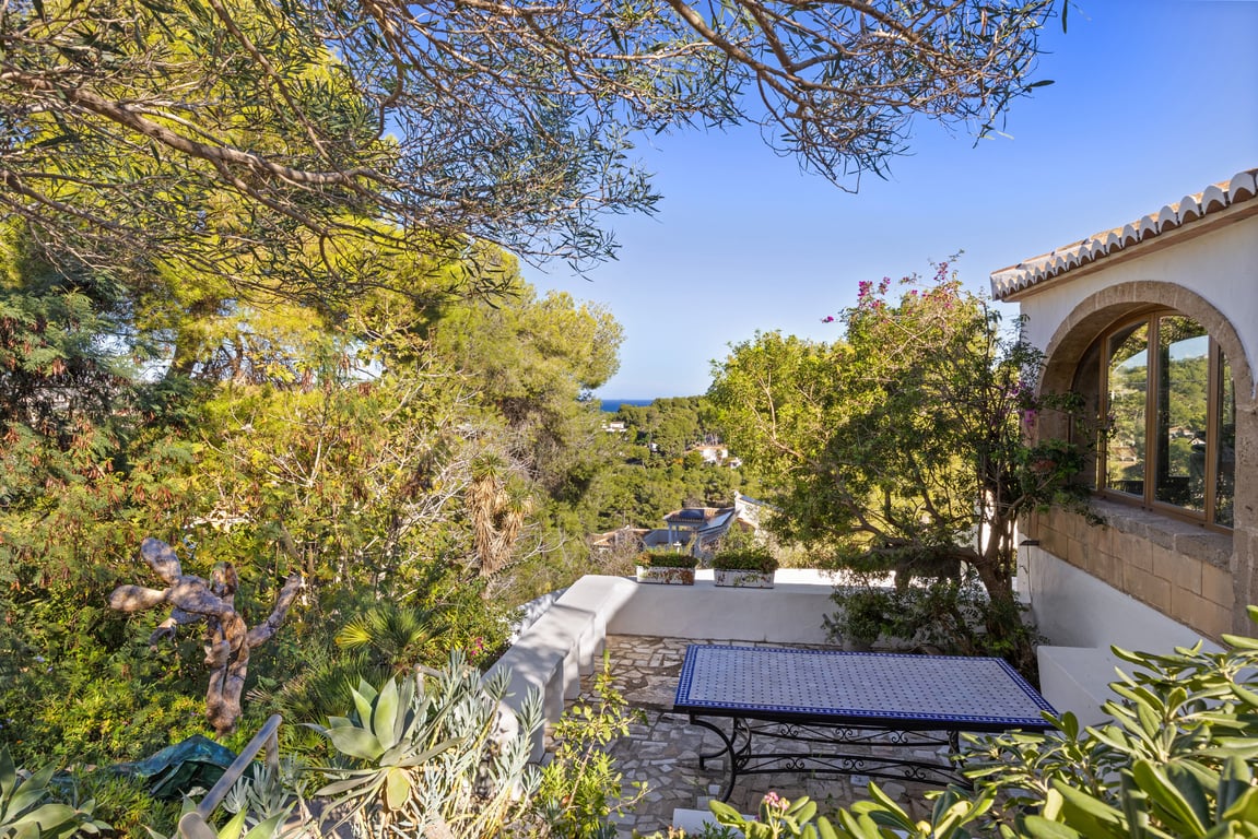 Außenterrasse mit Natursteinpflaster und Mosaiktisch, umgeben von mediterraner Vegetation mit Meerblick, der durch Pinien und traditionelle Architektur eingerahmt wird.