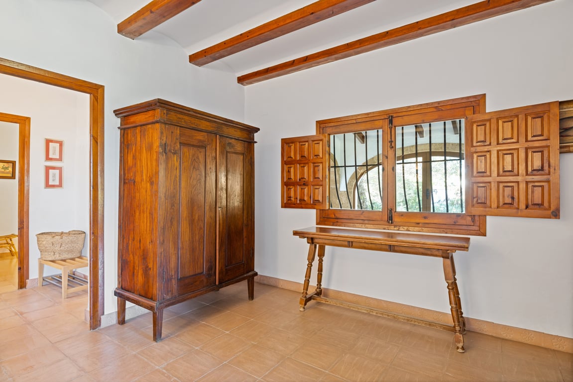Eingangsbereich mit sichtbaren Holzbalken, handgefertigten Keramikfliesen und rustikalen Möbeln. Das Fenster bietet traditionelle Fensterläden und Sicherheitsgitter.