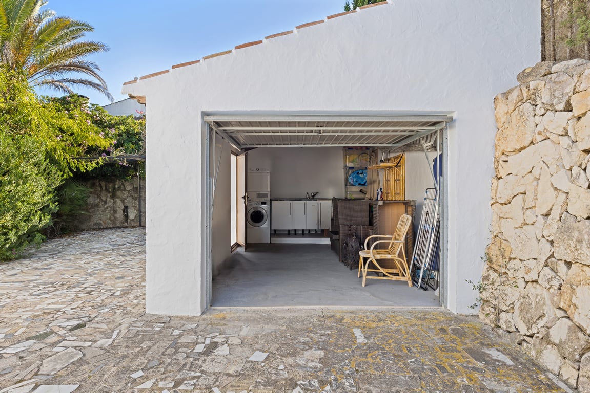 Funktionale Garage mit Hauswirtschaftsbereich, ausgestattet mit Waschmaschine, Trockner und Schränken, neben einer Natursteinmauer und einer gepflasterten Einfahrt.