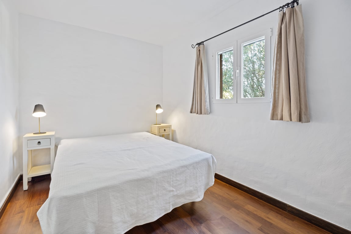 Helles Schlafzimmer mit Holzboden, weißen Wänden und doppelt verglastem Fenster mit Blick ins Grüne. Inklusive Nachttischen und funktionalen Leselampen.