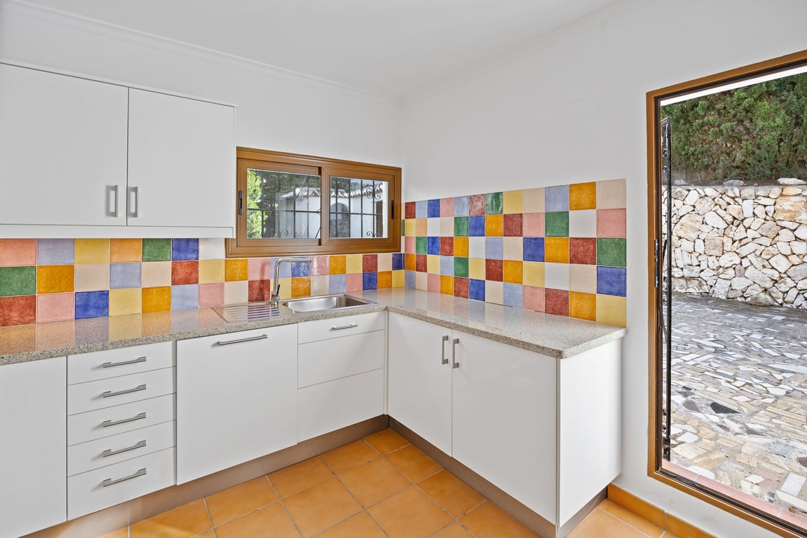 Küche mit weißen Fronten, Granitarbeitsplatte und einem markanten bunten Fliesenspiegel. Ausgestattet mit Terrakottaboden und direktem Ausgang zum gepflasterten Außenbereich.