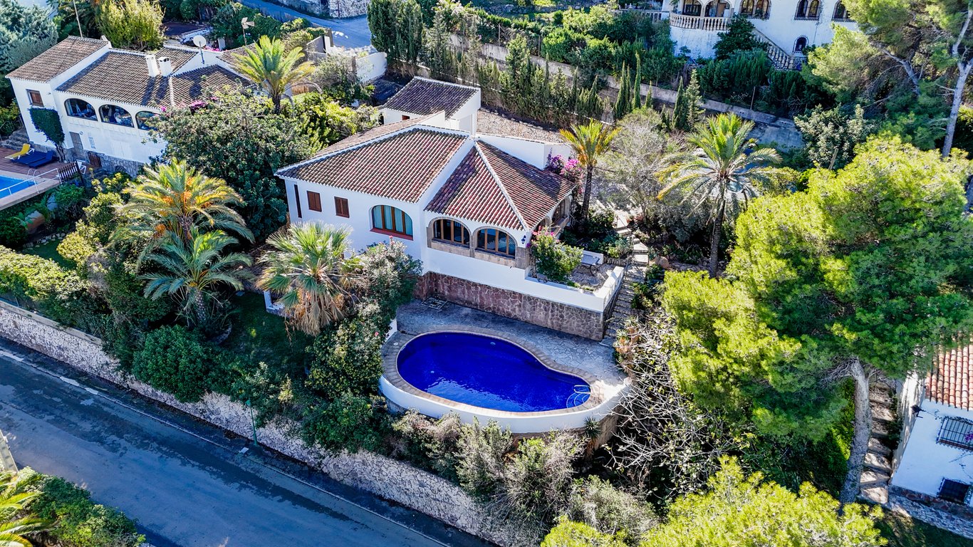 Luftaufnahme der traditionellen Villa mit nierenförmigem Pool, Terrakottadach und bogenförmiger Naya, eingebettet in einen mediterranen Garten mit Palmen.