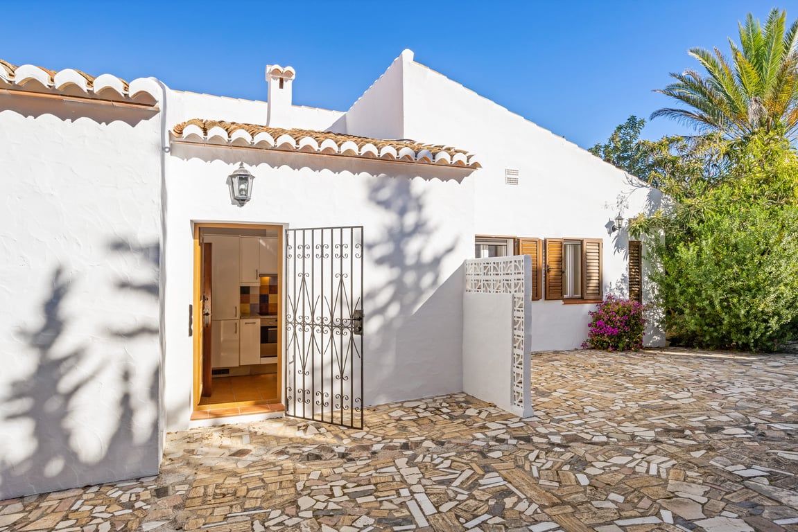 Mediterrane Außenansicht mit weiß getünchten Wänden, Natursteinpflaster und einem Nebeneingang mit Schmiedeeisengitter sowie traditionellen Holzfensterläden.
