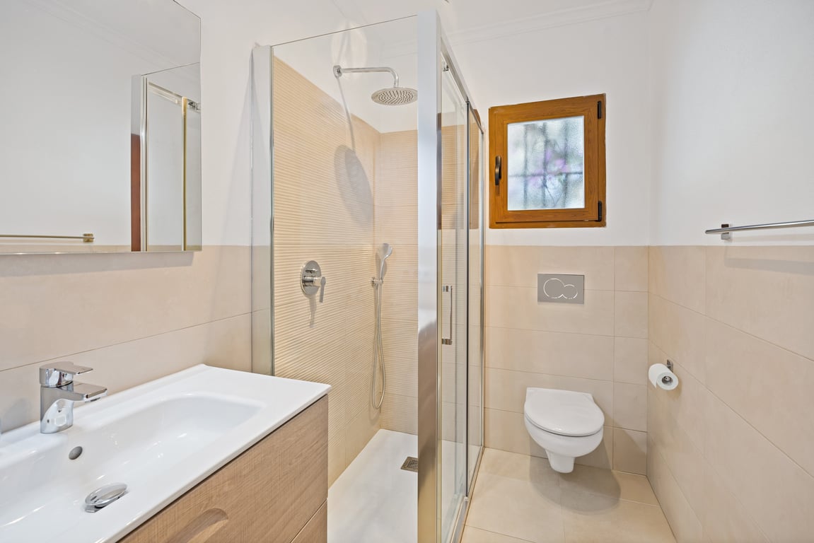 Modernisiertes Badezimmer mit Glasduschkabine, Waschtisch aus hellem Holz und neutralen beigefarbenen Fliesen an Wänden und Boden.